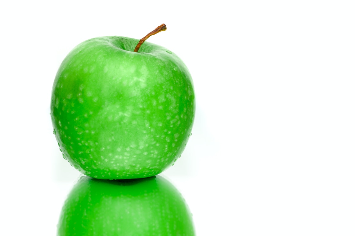 Grüner Apfel mit Wassertropfen auf glänzender Oberfläche vor weißem Hintergrund.