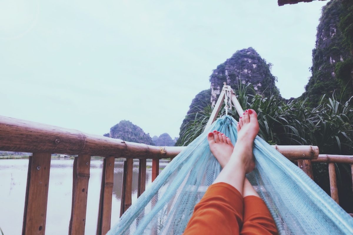 Person entspannt sich in einer Hängematte mit ausgestreckten Füßen, vor einer tropischen Kulisse aus üppigem Grün und fernen Kalksteinbergen.